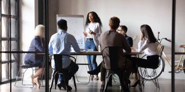 Diverse workers listening african businesswoman tell about strategy at meeting Six diverse businesspeople gathered in modern board room, different age ethnicity workers listen african woman coach tell about strategy, reporting using flip chart. Training and negotiations concept